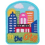 A sunny cityscape sits on top of a green hill. The words In The City are embroidered below.