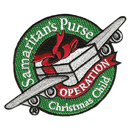 A shoe box with airplane wings is surrounded by a red and green ribbon that reads Samaritan's Purse, Operation Christmas Child.