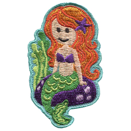 A mermaid sits on a rock at the bottom of the ocean floor. She has long hair with a starfish, two dots for eyes, and a U shaped smile. Two stocks of sea weed sway off to the left of the crest.