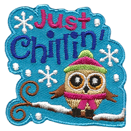 A little owl is bundled up in a toque and scarf as it sits on a snow covered tree branch. Snowflakes fall from the sky accompanied by the text 'Just Chillin'.
